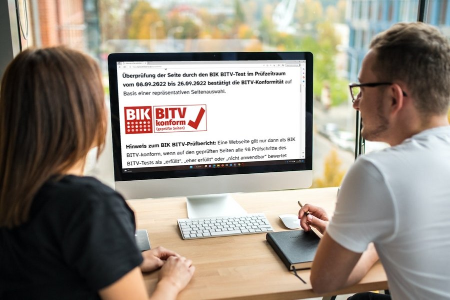 Großansicht Zwei Personen sitzen vor einem Monitor auf dem ein Ausschnitt aus der Erklärung zur Barrierefreiheit mit BIK BITV-Test Zertifikat zu sehen ist.