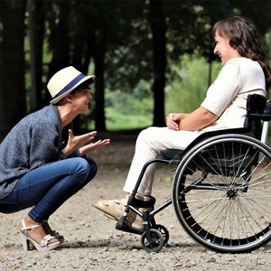 Eine Frau sitzt im Rollstuhl, eine andere hockt vor ihr. Beide unterhalten sich und lachen.