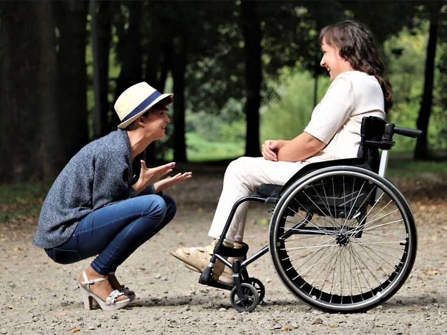Großansicht Eine Frau sitzt im Rollstuhl, eine andere hockt vor ihr. Beide unterhalten sich und lachen.
