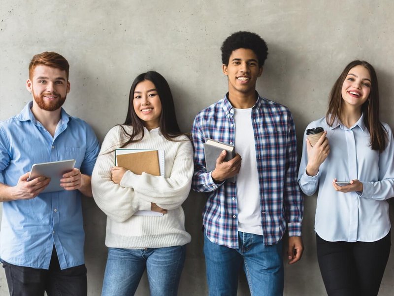 Vier junge Leute lehnen entspannt an einer Wand und lächeln in die Kamera