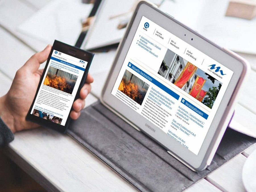 Großansicht Eine Hand hält ein Handy, daneben auf dem Schreibtisch steht ein Tablet, beide zeigen Ansichten der barrierefreien Webseite der Stadt Meerbusch.